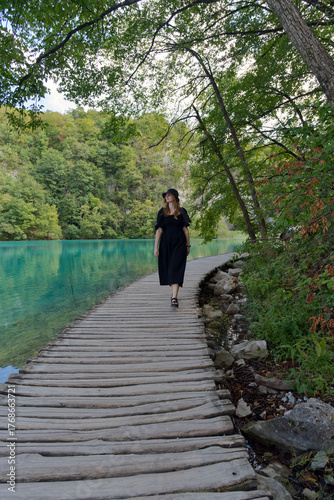 A girl strolls peacefully along a rustic boardwalk beside Plitvice Lake’s turquoise waters, surrounded by lush foliage and serene reflections, embracing nature’s calm in a moment of quiet solitude.