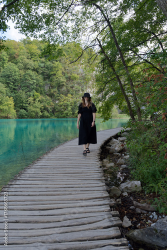 A girl strolls peacefully along a curved wooden boardwalk beside Plitvice Lake’s turquoise waters, surrounded by lush greenery and calm reflections, capturing the essence of nature’s serenity