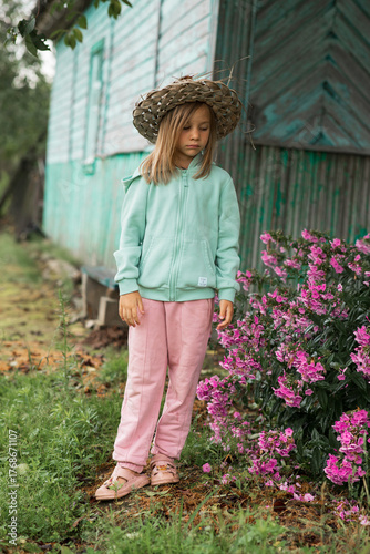 a blonde girl wearing a straw hat is looking at beautiful pink flowers in the garden