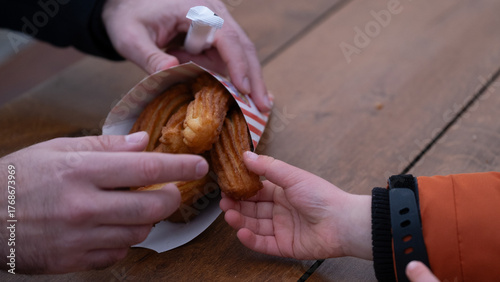 Close-up of adult and child hands sharing a paper bag of churros on a wooden table outdoor