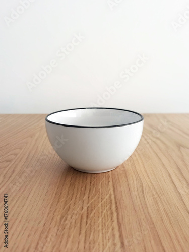 Close-up of a white ceramic bowl on a wooden background and a white wall. Front view of an elegant, minimalist bowl. Ceramic household utensils. Close-up of a white stoneware mug, simple design.