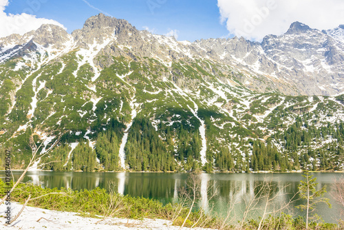 alpine lake in spring on a sunny day. travel landscape of rysy ridge in high tatra mountains of poland. snow among rocks and spruce forest on the hillside reflection in water. blue sky with clouds