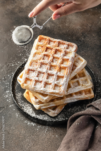 Delicious belgian waffles in stack on dark background. Woman hand is sprinkling waffles with powdered sugar close up