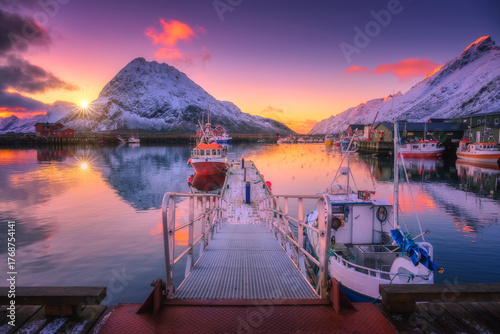 Fishing boats in harbor at sunset with snowy mountains and colorful sky in Lofoten Islands, Norway. Scenic waterfront with pier, vessels, reflection in calm water, and Arctic winter landscape
