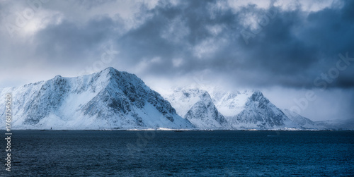 Majestic snow-capped mountains rise from the deep blue ocean under dramatic cloudy sky. Winter landscape with snowy rocks, overcast sky and sea in the Lofoten islands, Norway. Nature. Arctic scenery