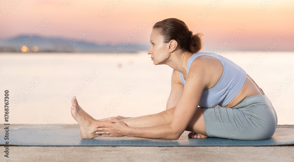 Fototapeta premium Young slender woman practices yoga on deserted seashore doing asanas, performs physical exercises. During sunset, girl performs variations of Janu Shirshasana
