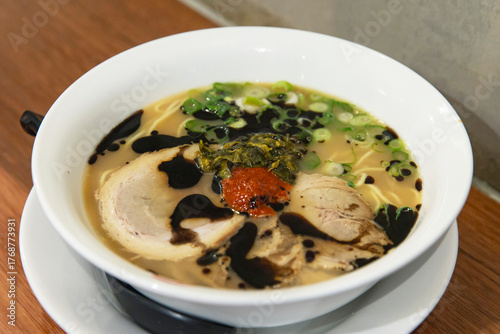 Bowl of rich pork tonkotsu ramen with black garlic oil, spicy paste, pickled greens and green onions in savory broth on wooden table