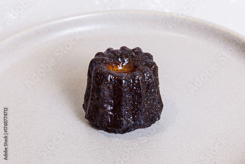 Single canelé pastry on a light ceramic plate, rich dark caramelized crust with glossy sheen, minimal presentation highlighting texture