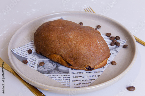 Coffee bun with golden crust served on a ceramic plate, surrounded by scattered coffee beans, presented on vintage newspaper lining