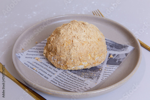 Round dome-shaped cake covered in fine beige crumbs, served on neutral ceramic plate lined with printed paper, showcasing soft texture