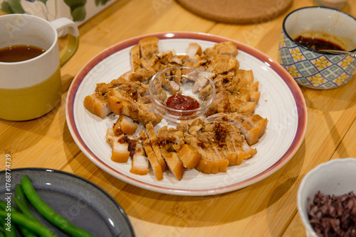 Home dining setup with sliced pork belly in circular arrangement, spicy dipping sauce in center, surrounded by cups and side dishes on wooden table