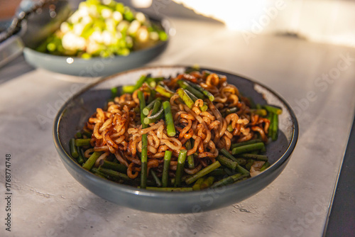 Closeup of stir fried noodles with scallions in ceramic bowl, glistening under warm light in kitchen setting, blurred bowl of vegetables in background
