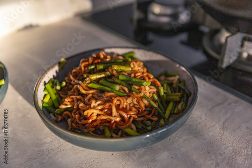 Bowl of spicy stir fried noodles with vibrant green vegetables, placed on kitchen counter under warm natural light with visible gas stove background