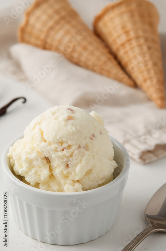 Vanilla Bean Ice Cream Scoop in White Ramekin with Waffle Cones, Vanilla Pods, and Spoon on White Surface