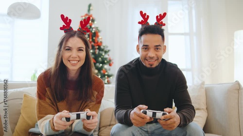 Happy multiracial couple wears reindeer antlers while playing video games together in their living room, celebrating Christmas and New Year with festive decor around them.