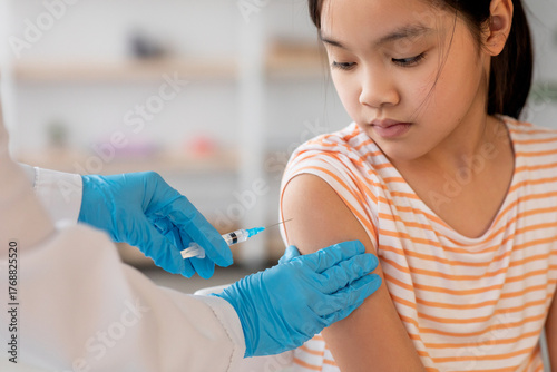 A young girl sits calmly while a healthcare worker administers a vaccine to her arm. The clinic is well-lit and organized, focusing on child healthcare.