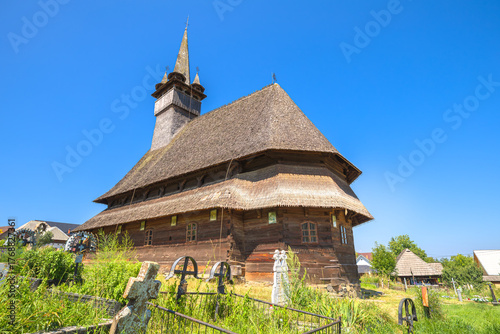 Historic wooden Maramures church of Saint Nicholas in Budesti Josani, Romania, standing under a clear blue sky, surrounded by green grass and traditional cemetery crosses. Maramures of 1600s