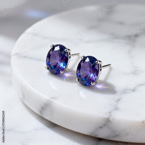 A stunning pair of stud earrings featuring large oval-cut tanzanite gemstones displaying rich blue and purple hues is beautifully presented on a round white marble display surface.