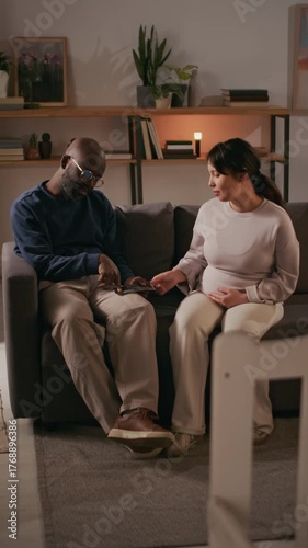 Vertical shot of excited interracial couple viewing ultrasound photo of their upcoming baby while sitting on couch in their cozy nursery