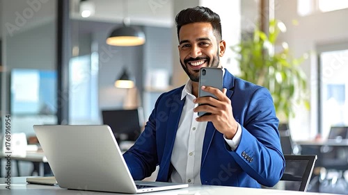 Smiling man in a suit holds a phone near a laptop. Office setting. Natural light