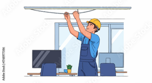 A male electrician in a yellow hard hat and blue overalls works on wiring a ceiling light fixture in an office environment with computers and desks.