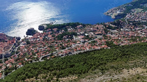 Aerial wide-angle drone faces outward over Dubrovnik’s Adriatic coastline, capturing terracotta rooftops, rocky shores, and the deep blue sea sparkling under bright sunlight along the coastline