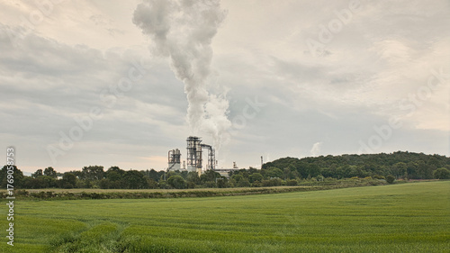 a landscape view and across the green fields there are industrial buildings letting smoke off into the atmosphere from the chimney