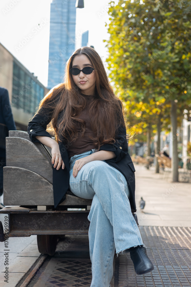 Fototapeta premium Fashion-forward young Asian woman wearing sunglasses, blazer and denim jeans, sitting on an industrial-style bench in a modern city street. Urban lifestyle, street fashion and contemporary vibe