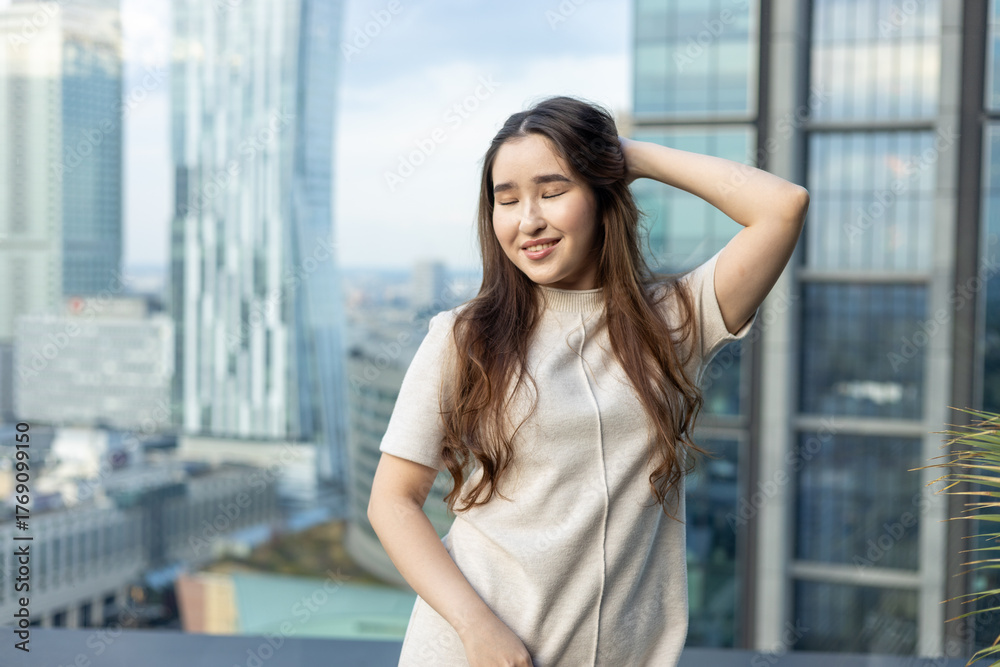 Fototapeta premium Asian woman in chic beige dress holding a handbag on a rooftop terrace with city skyline — full-length fashion & lifestyle portrait