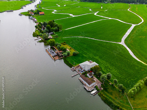 A peaceful river scene with boats sailing calmly past green fields, traditional houses, and a historic windmill, capturing the charm of the countryside and serene rural life.