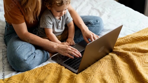 Parent and toddler using a laptop together on bed in cozy home setting, learning and bonding during remote work and online education time with family technology and support
