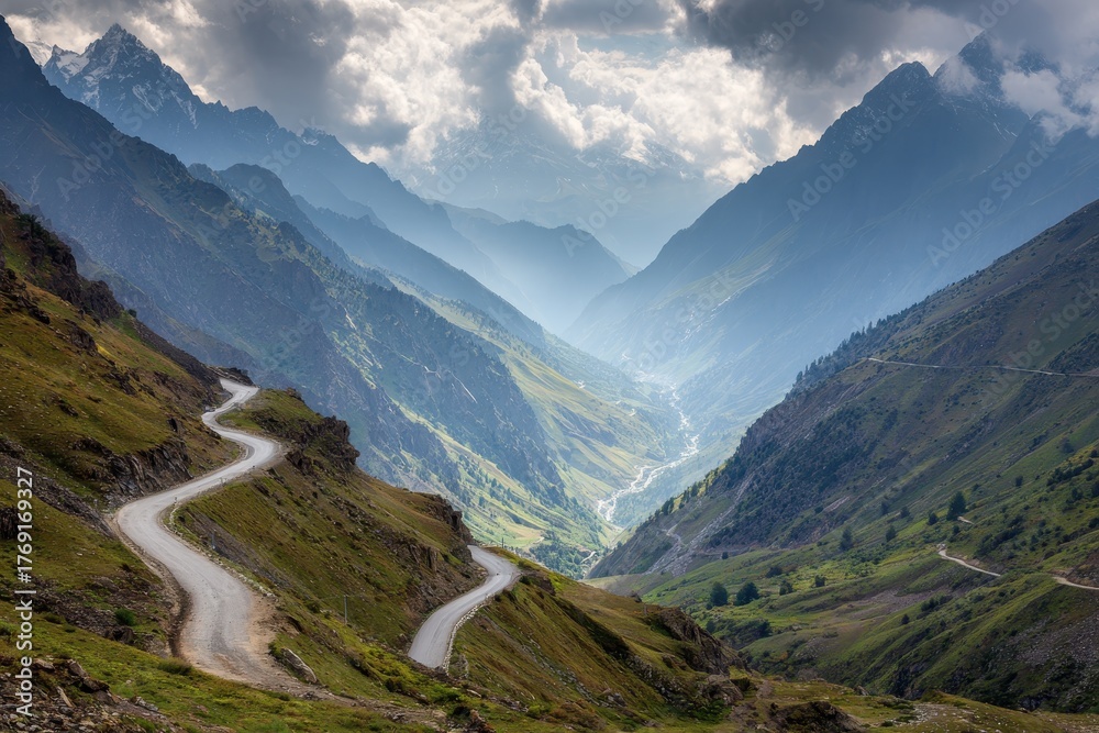 Obraz premium Winding mountain road descends into a breathtaking valley surrounded by majestic peaks and shrouded in atmospheric clouds during the afternoon light