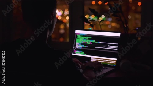 A young male programmer in front of a window with a lights of night city, on his laptop screen is code. out of focus. 