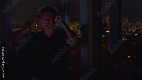 A young male programmer in front of a window with a lights of night city, on his laptop screen is code. out of focus. 