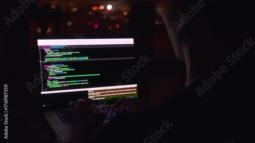 A young male programmer in front of a window with a lights of night city, on his laptop screen is code. out of focus. 