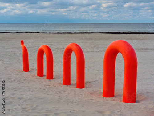 A row of bright orange, tube-like arches on a sandy beach,  the calm sea and cloudy sky.