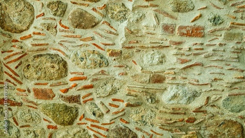  Close-up shot of an old stone and brick wall with a rough texture. The wall is made of various natural stones and fragments of orange bricks set in cement, creating a unique and rustic pattern