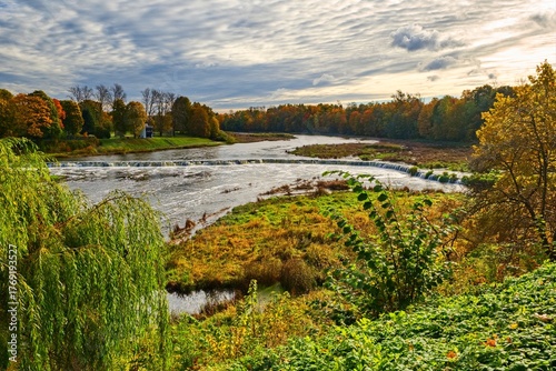 Picturesque autumn landscape with the Venta Rumba, Europe's widest waterfall, in Kuldiga, Latvia. The Venta River flows through dolomite rapids, surrounded by colorful autumn trees.