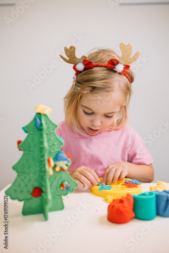 A young girl wearing festive antlers enjoys crafting clay decorations for a plastic Christmas tree