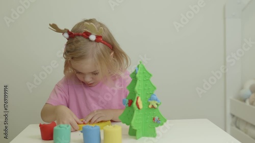 A young girl wearing festive antlers enjoys crafting clay decorations for a plastic Christmas tree