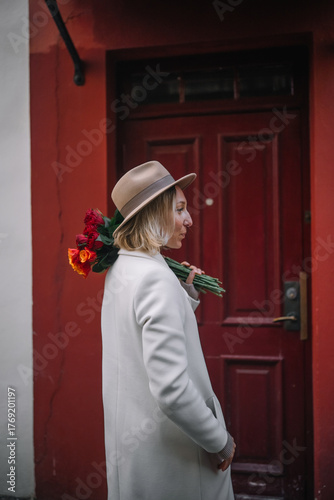 Woman holding flowers walks near red door in urban setting during daytime