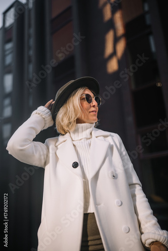 Stylish woman in a white coat and hat poses confidently outdoors against a modern urban backdrop