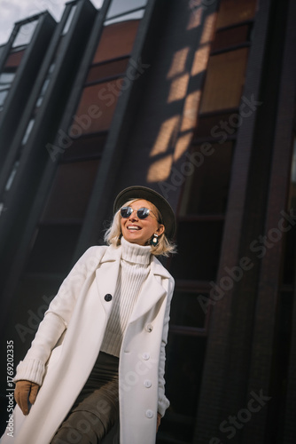 Stylish woman in a white coat and hat poses confidently outdoors against a modern urban backdrop