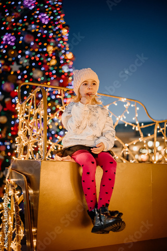 Child enjoying festive treats on a decorated sleigh amidst bright holiday lights