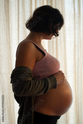 Portrait of Pregnant Woman in Backlight with a Curtain as a Background. Pregnancy Shooting in Home Interior