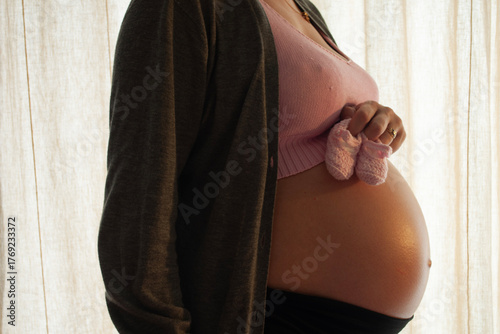 Detail of a Pregnant Woman Holding Pink Handmade Baby Shoes on Her Belly. Pregnancy Shooting in Home Interior