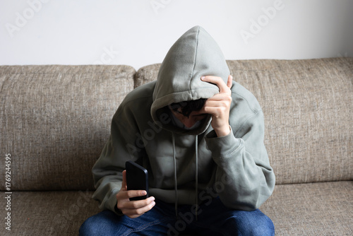 Cyberbullying, negative in social media online issue in teenager. Upset teen boy sitting near window using smartphone at home. 