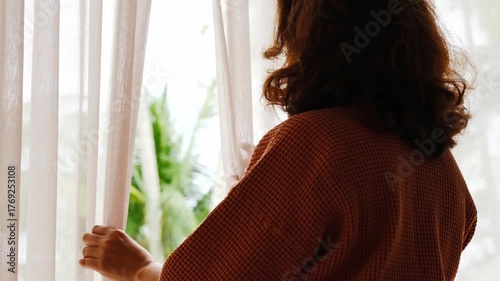 The woman was at the window in the bedroom. She opened the curtains on the window. In the morning and she looks at the view of mountains and trees at sunrise.