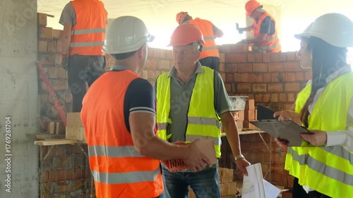 Worker discusses processes with foreman at construction site. Specialist holds brick in hands and stands near engineer against wall