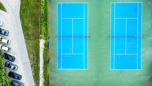 Blue tennis courts from above, showing nets and white lines, surrounded by green landscaping and an adjacent car parking lot with vehicles